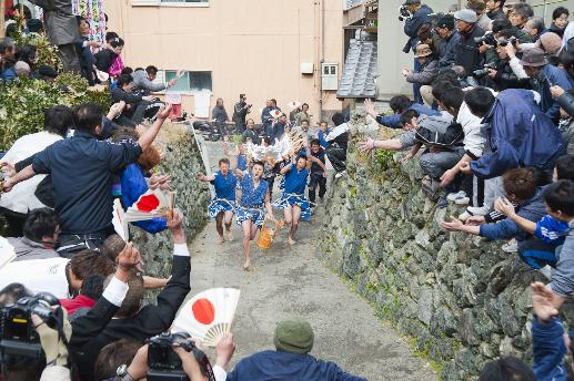 両側の石垣の上の、日の丸の模様の扇子を持った大勢の人々が、石垣に囲まれた小道を上ってくる若者たちを待ち構えている様子の写真