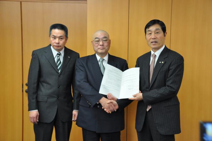 男性3名が並び、中央と右側の男性が協定書を一緒に持ちながら握手をしている写真
