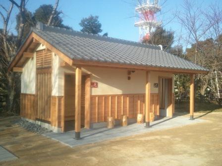 周囲に木々があり、瓦屋根と壁の一部が杉で囲われているのが特徴的な、和風デザインの城山公園トイレの写真