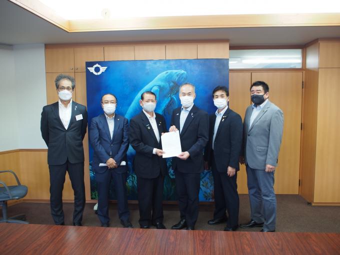 マスクをしている男性6人が横に並び真ん中2人が紙を持っている写真