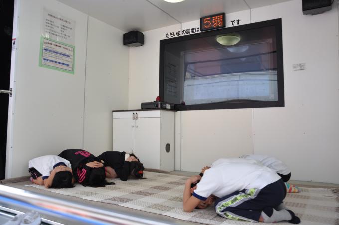 画像:地震体験車の中で、震度5弱の揺れを体験し、床に伏せて頭を手で抑え、揺れから身を守っている5人が写った写真