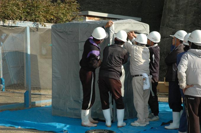 画像:白いヘルメットを被った数名が広場に敷かれたブルーシートの上に災害用の仮設トイレを組み立てている様子を写した写真