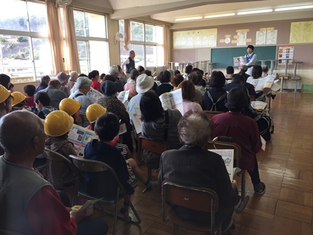 画像:学校の教室で、資料を持ち椅子に座って前を向いている数十人と黒板の前で資料を見せながらこちらに向かって説明をしている人が写っている写真