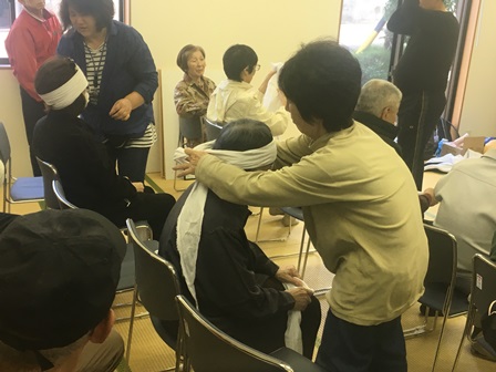 画像:椅子に座っている人の頭に包帯のようなものを巻きつけて手当ての訓練を行っている人など10名ほどが写っている写真