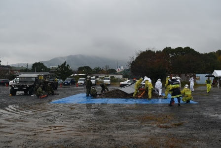 画像:雨が降っている中、広場のブルーシートの上に置かれたがれきの周りに10人ほどの人が集まって生き埋め者救出訓練をしている様子が写った写真