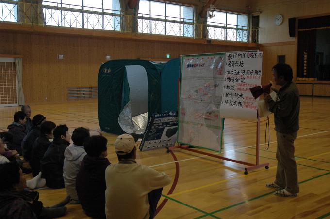 画像:体育館の中で、避難所の運営訓練の説明をしている1人と、それを座って聞いている数名が写った写真