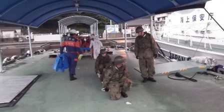 画像:船乗り場のような所で、迷彩服を着た人3名と消防隊のような制服を着た人数名が写っている写真