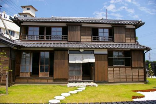 上に3つ下に1つの窓と掃き出し窓が1つあり、玄関には白い暖簾がかかっている昔ながらの木造2階建ての家の写真