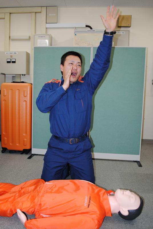 床に横たわる男性の人形を前に、手をあげ大声で助けを求める男性の写真