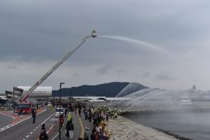 沿岸脇の道路にてはしご消防車のはしごを伸ばして上から放水する様子と、地上にて放水作業をする様子、それらを観覧する人々の写真。