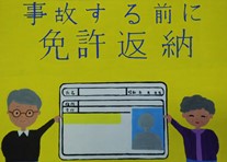 黄色い背景に大きな文字で「事故する前に免許返納」、真ん中に大きな免許証とそれを指さしている男性と女性が描かれたポスター作品