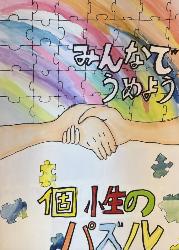 背景がパズルになっています。そこに握手をしている様子が描かれています。標語は「みんなでうめよう個性のパズル」です。