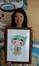 とば子育て応援キャラクターに選ばれた辻明日美さんが描いたキャラクターの絵を持ち、笑顔で立っている写真