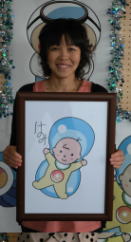 最終候補に選ばれた清水恵さんが自分の描いたキャラクターの絵を持ち、笑顔で立っている写真