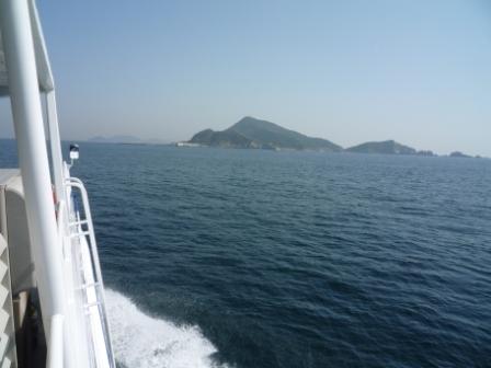 白いボート船が左端に写り、水しぶきを上げて遠くに見える島に向かっている様子の写真