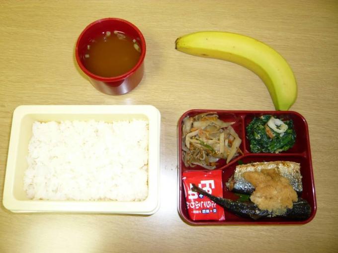机に並べられた配食サービス弁当（白飯と魚や煮物など3種のおかず弁当と、バナナと味噌汁）の写真