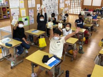 小学校の教室でマスク着用の児童が、もらったシェルレーヌを掲げて喜んでいる様子の写真