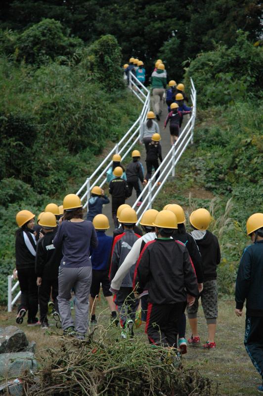 画像：訓練に参加している住民が、黄色いヘルメットを被り、整備された避難路を使って高台へ避難するために坂を登っている様子を後ろから写した写真