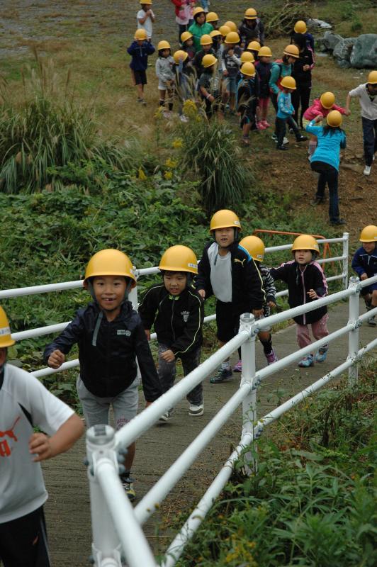 画像：訓練に参加している子供たちが、黄色いヘルメットを被り、整備された避難路を使って高台へ避難するために坂を登っている様子を正面から写した写真