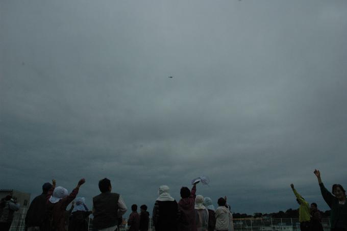 画像：曇っている空に小さく見えるヘリコプターと、それを地上から見上げて手を振っている人達が写った写真