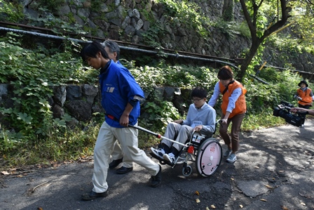 画像：石垣の前の登り坂を、男性が乗った車椅子を引っ張ったり後ろから押したりしている3人とそれを撮影しているカメラを持った人の手が写っている写真