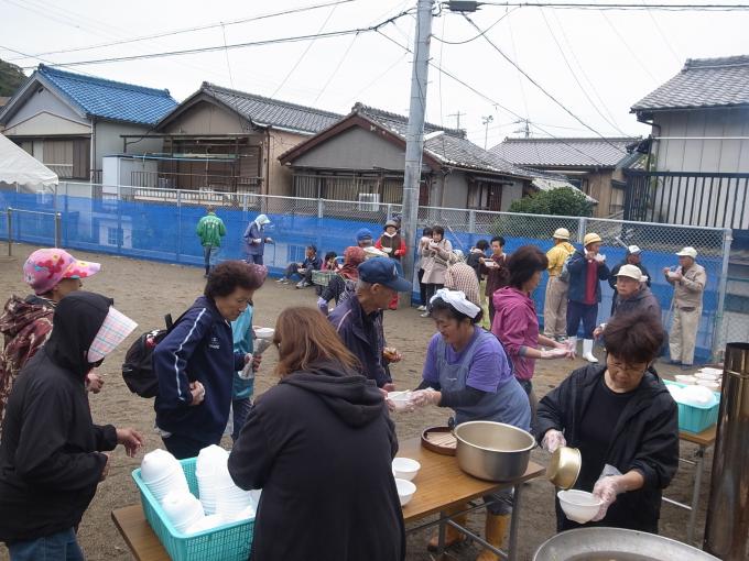 画像：炊き出しを器に盛り付けている人、それを受け取る人、受け取った炊き出しを食べている人が25人程写った写真