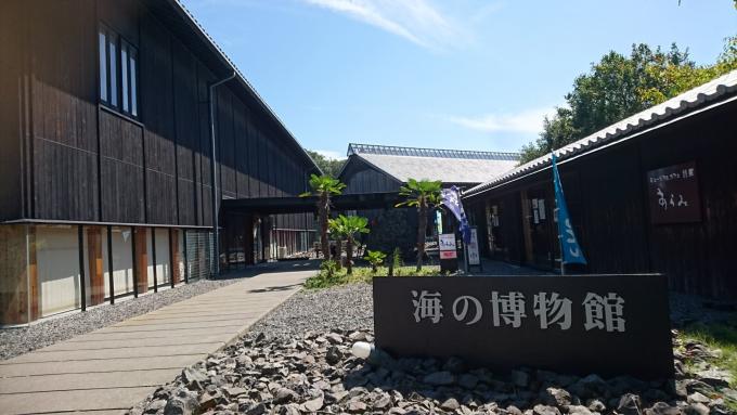 中央右寄りに白字で海の博物館と書かれた横長の石看板、砂利の上に石畳の道、その両脇に黒っぽい建物がある風景の写真