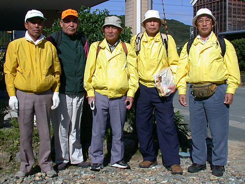 1人は緑、4人は黄色のジャンパーを着て帽子を被ったご年配の男性5人が横一列に並んで立っている写真
