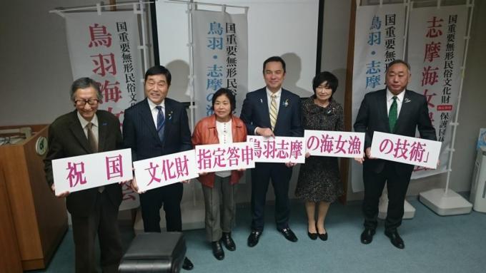 男女6人が白い背景に赤字で「祝国文化財指定答申鳥羽志摩の海女漁の技術」と書かれたパネルを持って立っている写真