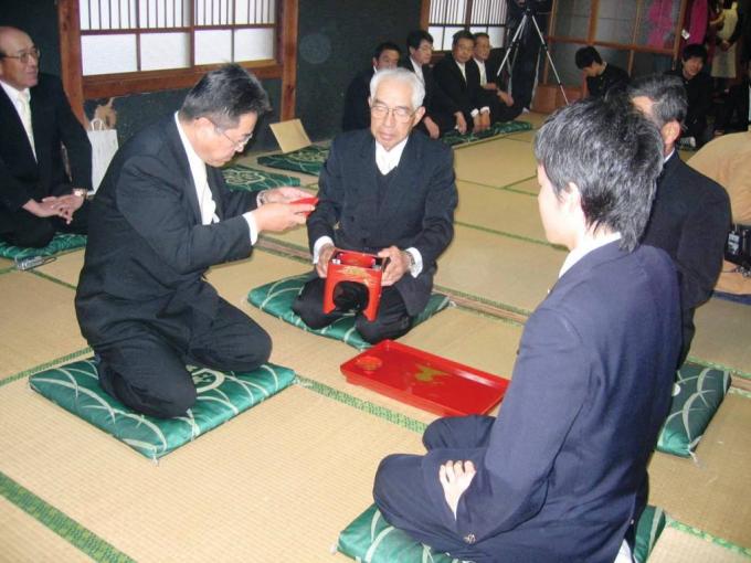 手前に正座をした男性が二人、奥に盆のようなものを持った男性が座っており、一人の男性がお酒のようなものをいただいている松尾の親取り子取りの写真画像