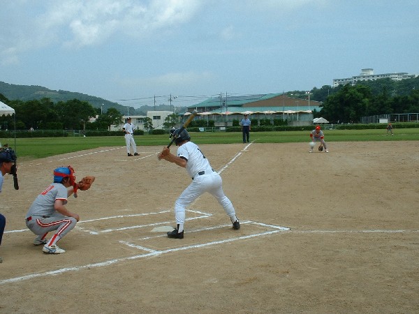 多目的グラウンドで行われる野球の試合で、打者がバットを持ち、ボールを待ち構えている写真