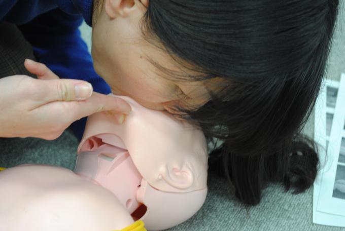 床に横たわる乳児の顎を2本の指で持ち上げ、乳児の口と鼻から息を吹き込む女性を近くから撮影した写真