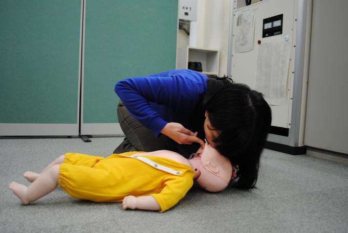 床に横たわる乳児の顎を2本の指で持ち上げ、乳児の口と鼻から息を吹き込む女性の写真