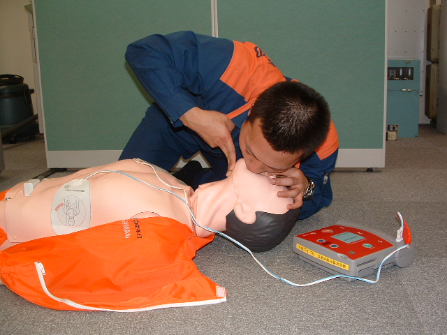 床に横たわりAEDの電極パッドをつけた男性の人形の顎に手をあて、口から息を吹き込む男性の写真