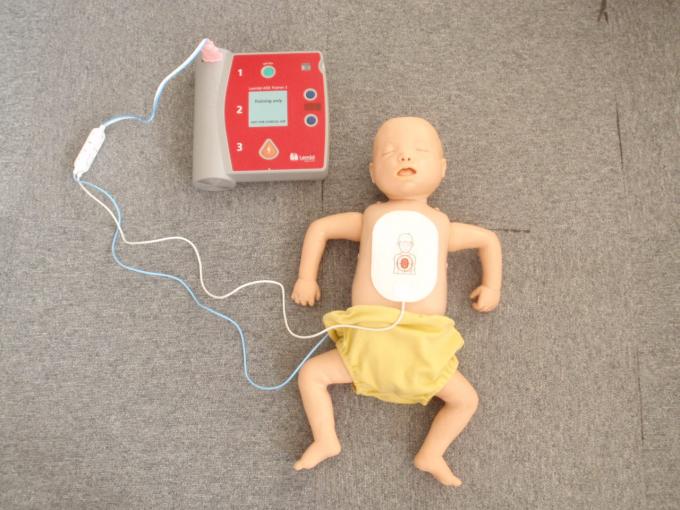 AEDから伸びる電極パッドを貼り付け、床に仰向けで横たわる上半身裸の乳児の人形の写真