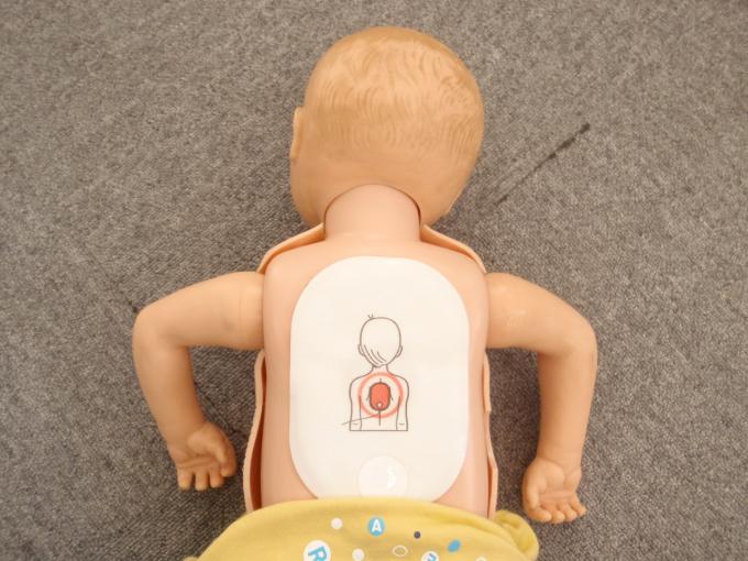 床にうつ伏せで横たわる上半身裸の乳児の人形に、イラスト入りのAEDの電極パッドを貼り付けた写真