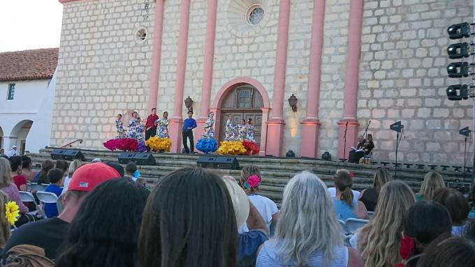 サンタバーバラ市の屋外のステージでカラフルな衣装を着て踊りを披露する人たちと、それを観覧する人たちの写真。
